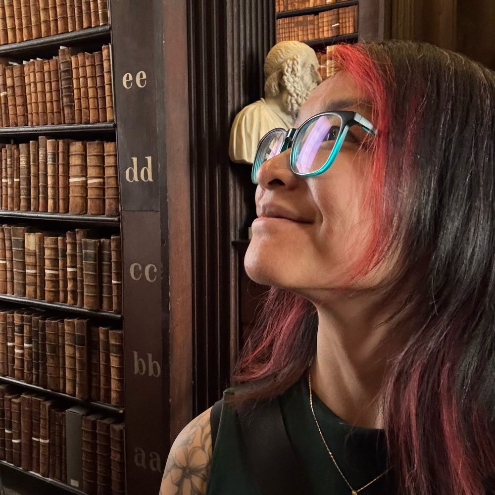 Profile photo of woman with red streaks dyed into her black hear, looking up and to the left against a backdrop of bookshelves with very old books